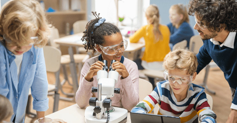 Young female student with safety goggles on sitting at a microscope, with fellow students nearby.