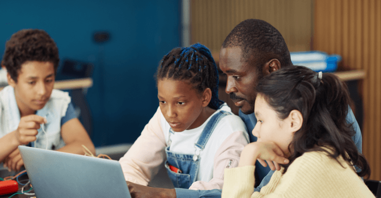 Two female students in front of a laptop with a Black male teacher sitting between them.