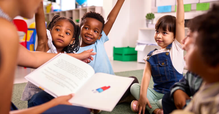 Group of four kindergarten age students gathered around a teacher who is reading a picture book. Three students are raising their hands.