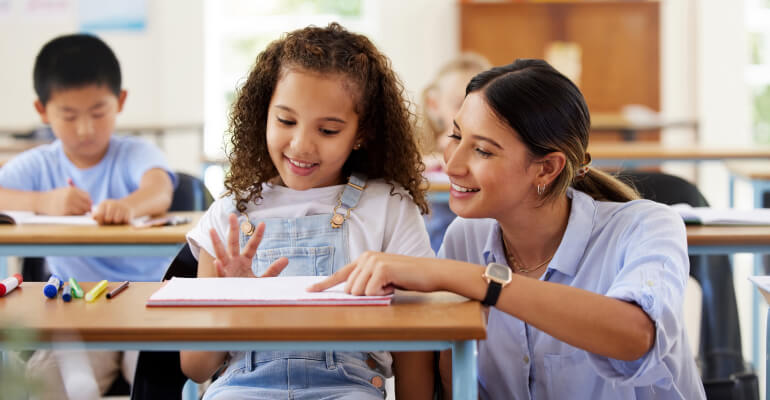 A female teacher crouched down beside a young fourth grade student's desk. The teacher points to a spot on an open book and both are smiling.