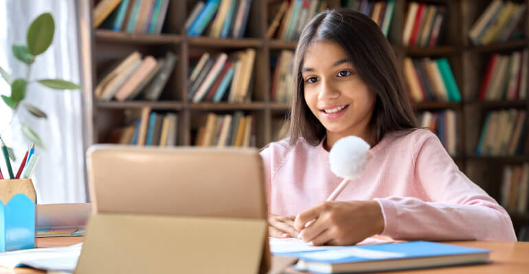 Upper elementary school girl student sits in front of a propped up tablet, holding a pencil and smiling.