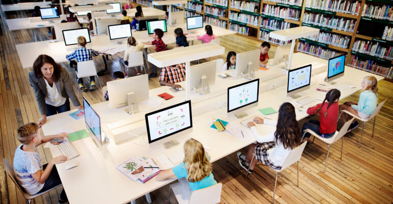 Overhead picture of a group of about a dozen elementary school students of different backgrounds sitting in a computer lab. They all work on projects while a teacher in a gray blazer leans against a table, looking at one student's computer screen.