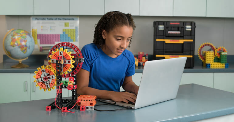 A middle school age female student in a blue shirt sits in front of a laptop with a robot she built as part of the Kid Spark coding lessons