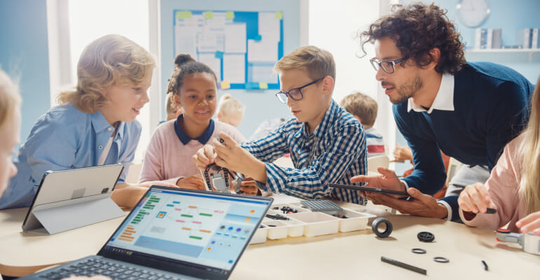 Male elementary school teacher leans across a table and talks with three elementary school students. A laptop is open in the foreground and gears and other building components are scattered on the table.