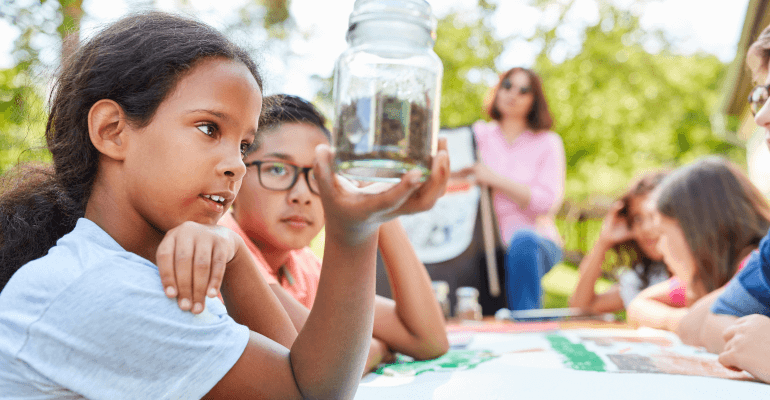 Young black female student holds up a mason jar with dirt in it while sitting outside. There are students and a teacher in the background.