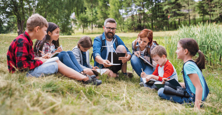 Middle aged man sitting outside with a group of schoolchildren. The man is holding a small windmill and smiling. He's teaching.