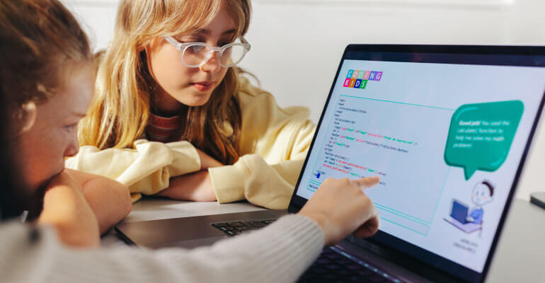 Two female students lean in toward a an open laptop. The student directly in front of the laptop is pointing at the screen where they are reviewing a coding lesson.