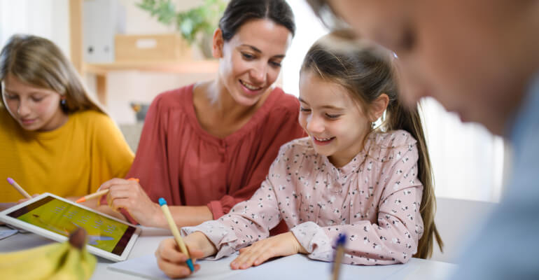 An elementary school girl student sits with a female teacher at a table. The girl holds a pencil and writes on a piece of paper while smiling. The teacher looks on, with a tablet sitting in front of both of them and another teacher in the background.