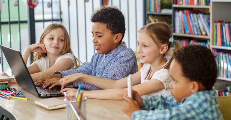 Group of four elementary school students sitting together at a large table. One student is typing at a laptop while the three others look on.