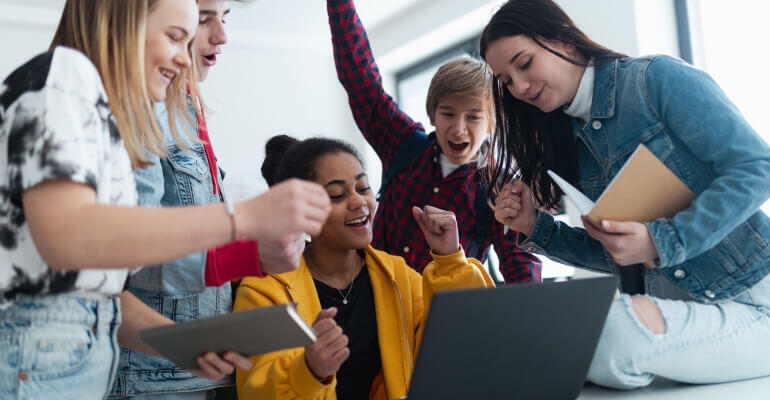 A group of five middle school students gather around a laptop. They are cheering on as they complete an assignment.