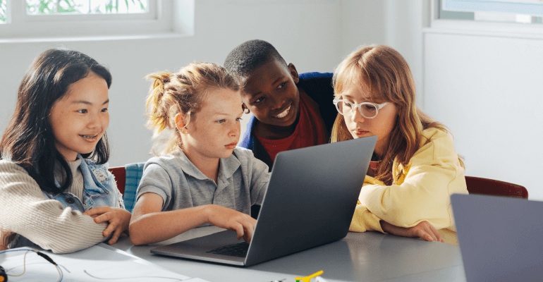 A group of elementary aged students of diverse backgrounds smile and look at a laptop.